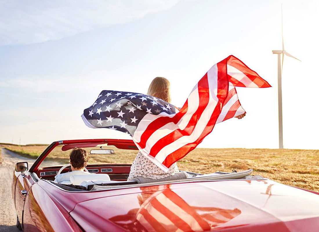 Insurance Solutions - Rear View of a Young Woman Standing Up in a Convertible While Holding Up an American Flag Next to her Husband Driving the Car While on a Road Trip