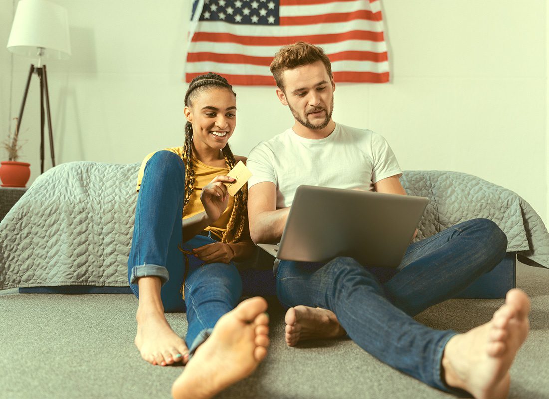 Service Center - Portrait of a Cheerful Young Couple Sitting Next to Their Bed in the Bedroom with an American Flag on the Wall While Using a Laptop